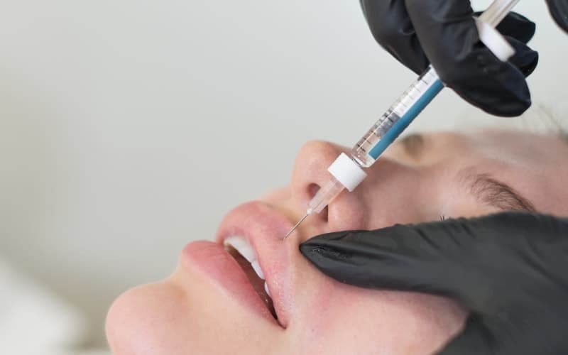 A clinician in black gloves administering a dermal filler injection to a patient's lip